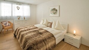 A bedroom with a bed, a nightstand, a chair, a dresser, a window with curtains, a ceiling light, and a wall-mounted picture. - Juggasunna Appartement/Fewo, Dusche und Bad, WC, 7-Bettwohnung by Interhome (Arosa)