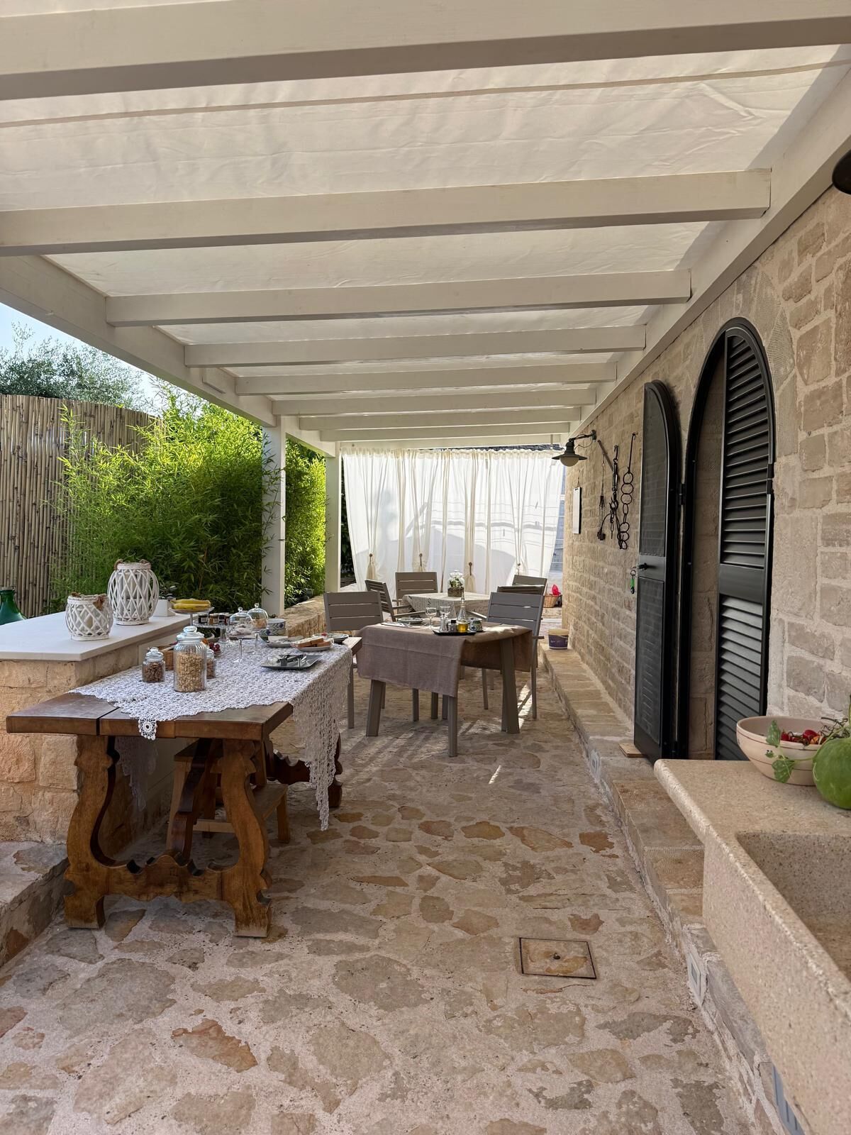 Un patio al aire libre con suelo de piedra, una mesa de madera lista para comer, y una zona cubierta con un toldo blanco.