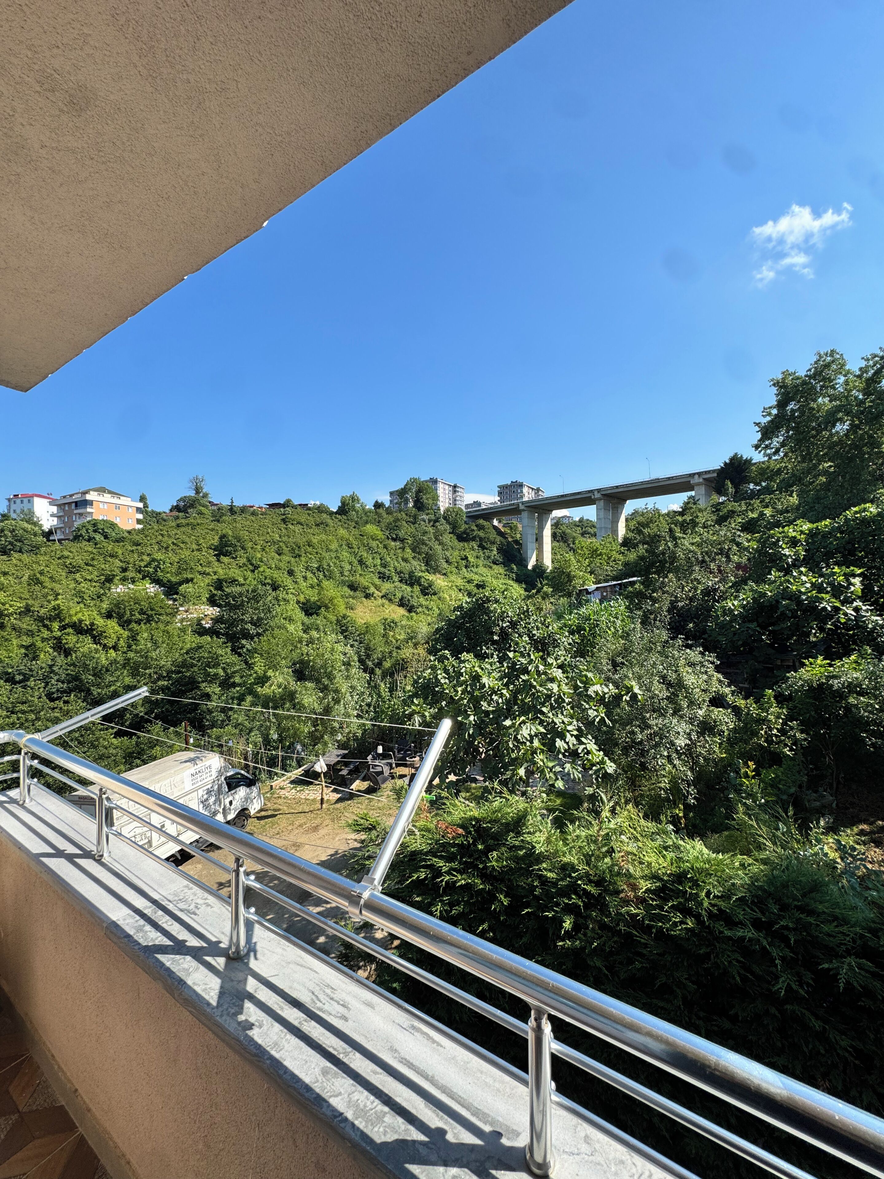 A balcony with a metal railing overlooking a hillside with dense greenery and a bridge in the distance.