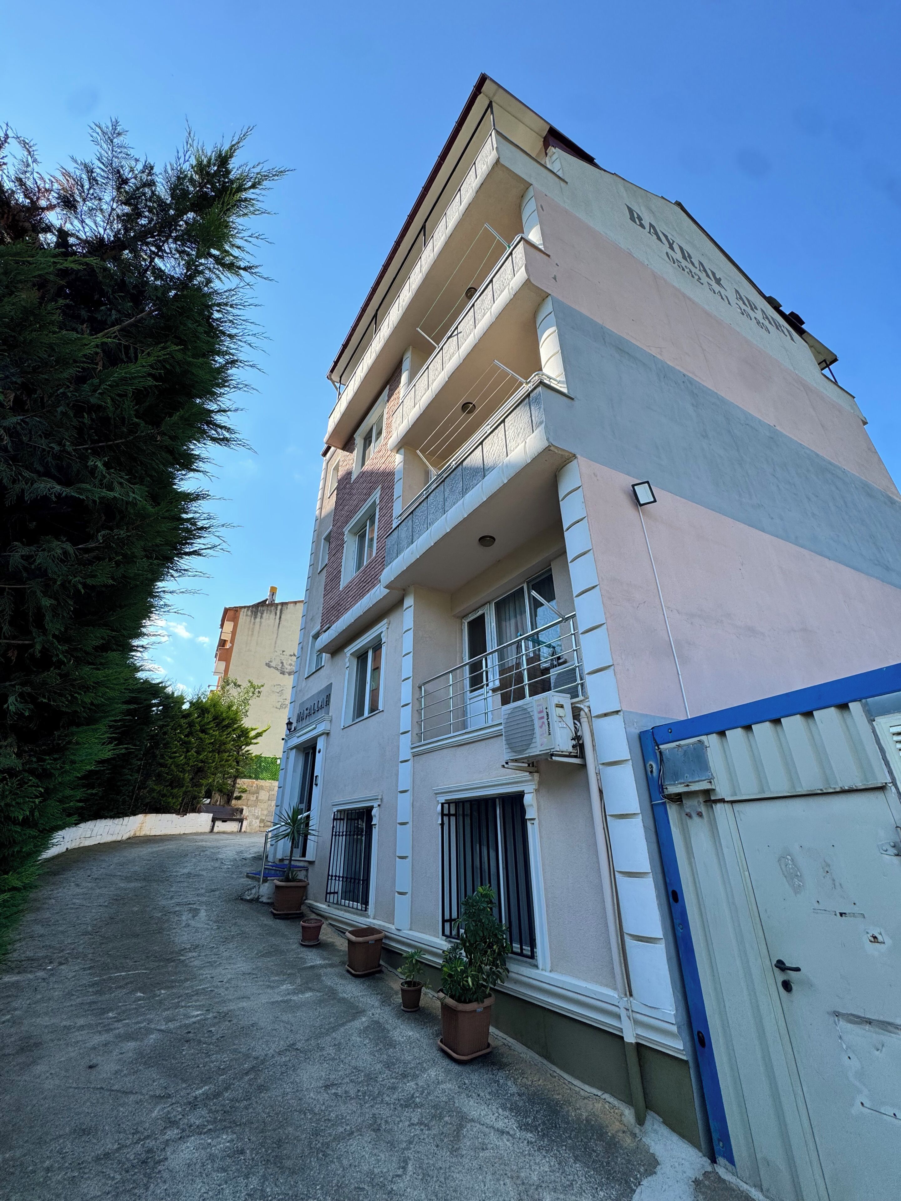 Un edificio de varios pisos con balcones, una entrada protegida por una reja y plantas en macetas a lo largo del camino.