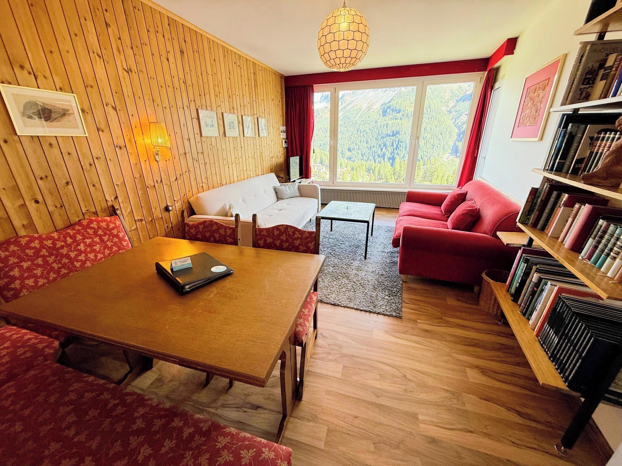 Ein gemütliches Wohnzimmer mit Holzwand, einem roten Sofa, einem roten Sessel, einem Holztisch und einem Bücherregal.