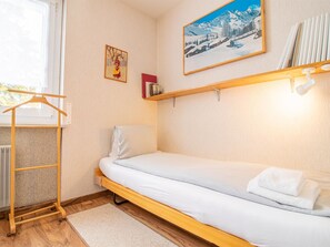 Ein Zimmer mit einem Einzelbett, einem Holzbettgestell, einem Heizkörper, einem Kleiderhaken und einem Regal mit Büchern. An der Wand hängt ein Bild von einer verschneiten Landschaft.