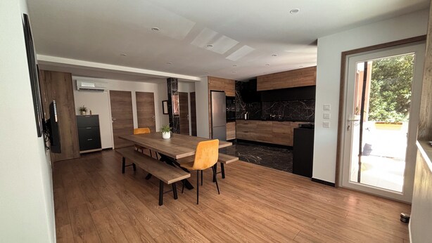 A modern kitchen with dark countertops, a wooden dining table with benches, and a sliding glass door leading to an outdoor area. - M&R Riviera • Near the beaches • Air Conditioning • Terrace • Modern 5-bedroom apartment • 8 people (La Seyne-sur-Mer)