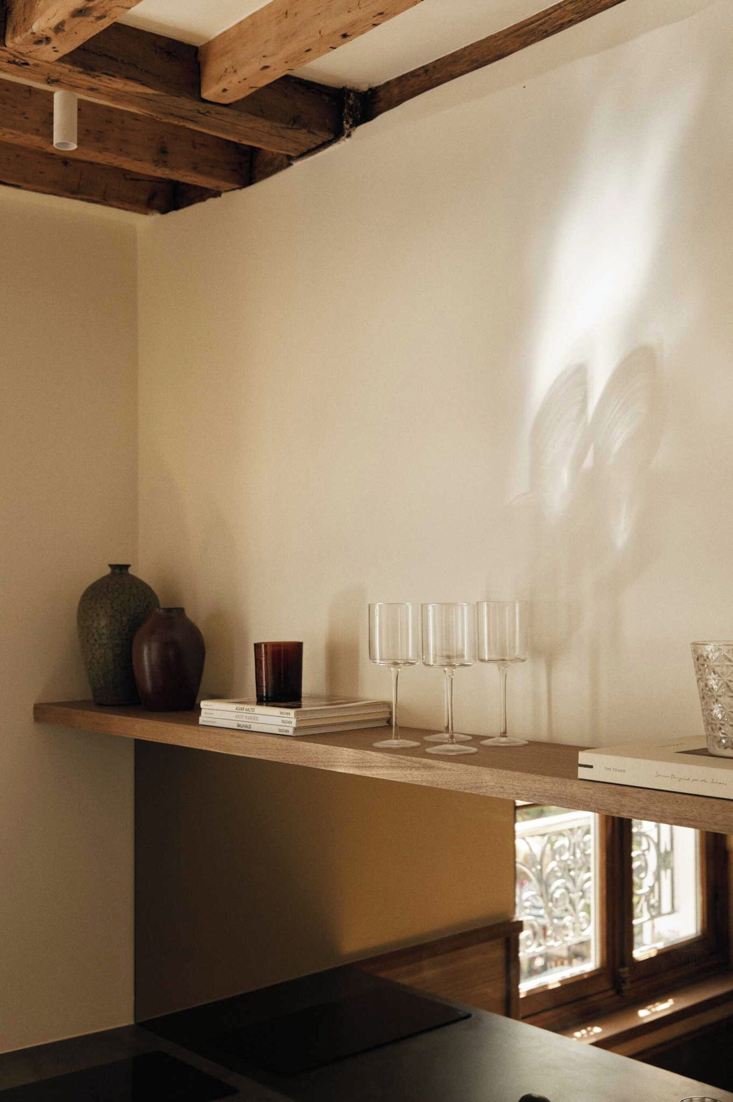 A room with a wooden ceiling, a window with a decorative iron grille, a shelf with books and a vase, and a wall-mounted light fixture.
