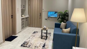 Una sala moderna con un sofá azul, una mesita auxiliar, una lámpara, un televisor montado en la pared y una zona de cocina con armarios y una encimera.