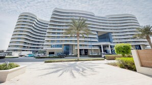 Un hôtel moderne à la façade incurvée, doté de nombreux balcons et d’un espace d’entrée aménagé avec des éléments paysagers.