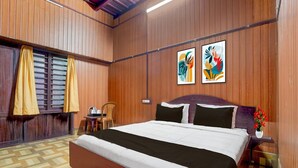 A room with a wooden floor, a bed with black and white bedding, a chair, a small table with a kettle, and two abstract paintings on the wall. - Deluxe Kallar Vattiyar Waterfalls Munnar (Devikolam)