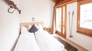 Een slaapkamer met een houten plafond, een bed met witte en donkere kussens, een klein nachtkastje en een raam met uitzicht op gebouwen.