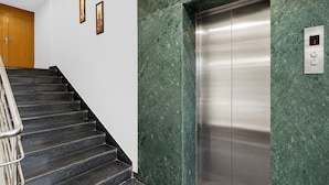 Een moderne lift met een metalen afwerking, naast een trap met donker marmeren treden en een metalen leuning.