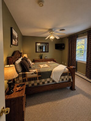 A bedroom with a bed, bedside table, lamp, mirror, ceiling fan, and a window with curtains.