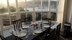 Une salle à manger aménagée dans une véranda, avec une table dressée pour quatre personnes, de grandes fenêtres et une vue sur un plan d’eau et des collines.
