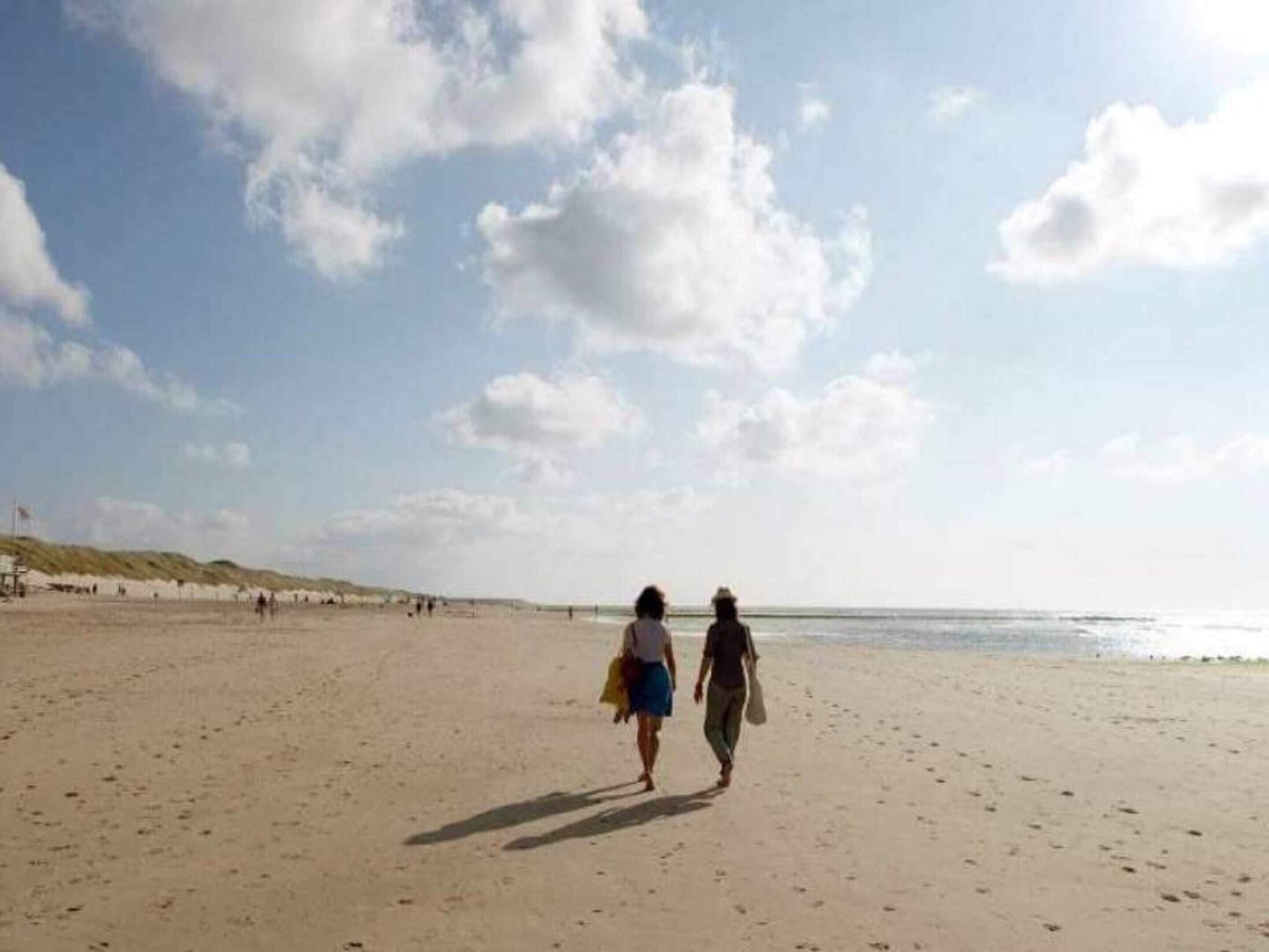 A wide sandy beach with people walking, a clear sky, and dunes in the background.