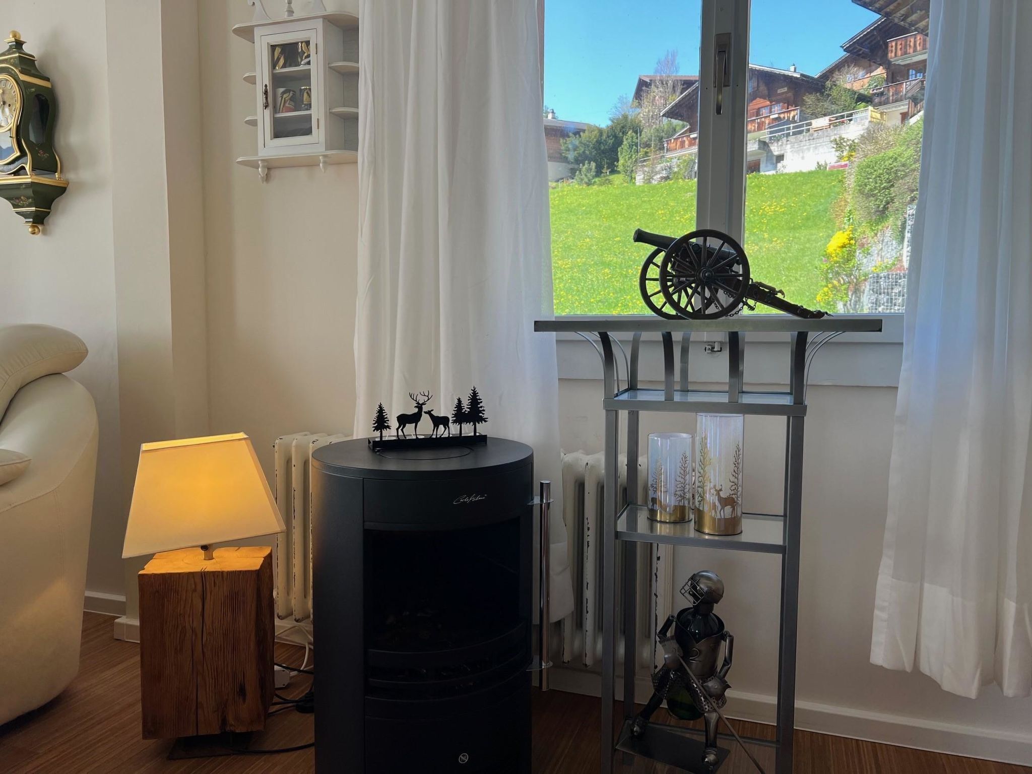 A room with a fireplace, a window with a view of houses, a lamp, and a shelf with decorative items.