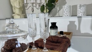 Un tocador de baño con una bañera, una rosa, dos vasos de cristal, una vela y un busto decorativo.