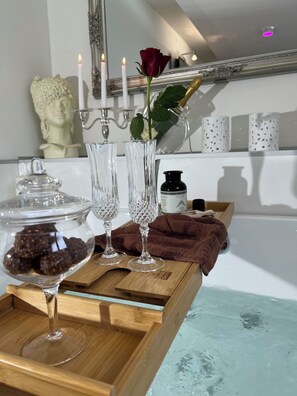 Ein Badezimmer-Waschtisch mit Badewanne, einer Rose, zwei Kristallgläsern, einer Kerze und einer Zierbüste.