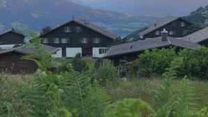 Een rij traditionele chalets met schuine daken, omringd door weelderige begroeiing en bergen op de achtergrond.