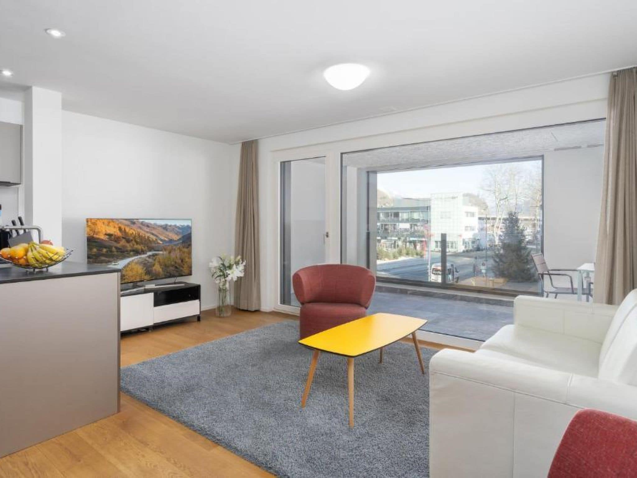 Un soggiorno moderno con un divano, una poltrona rossa, un tavolino, una TV e una finestra vista edifici e alberi.