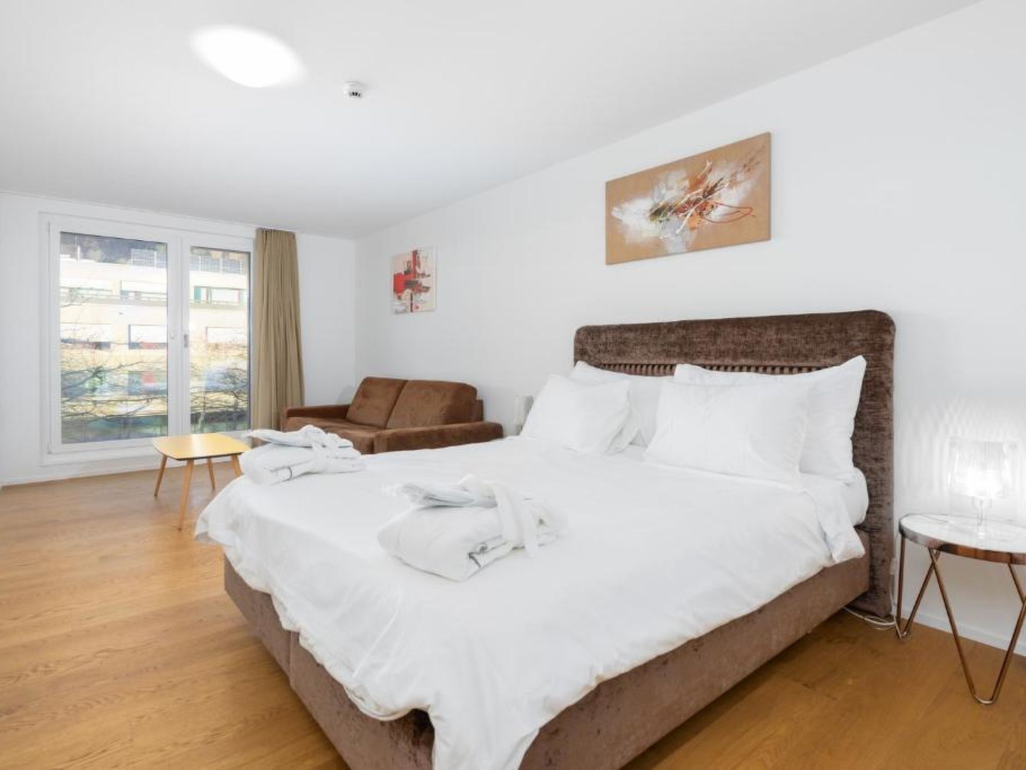 Habitación de hotel con una cama grande, cabecera marrón, una mesita, una silla y una ventana con vistas.