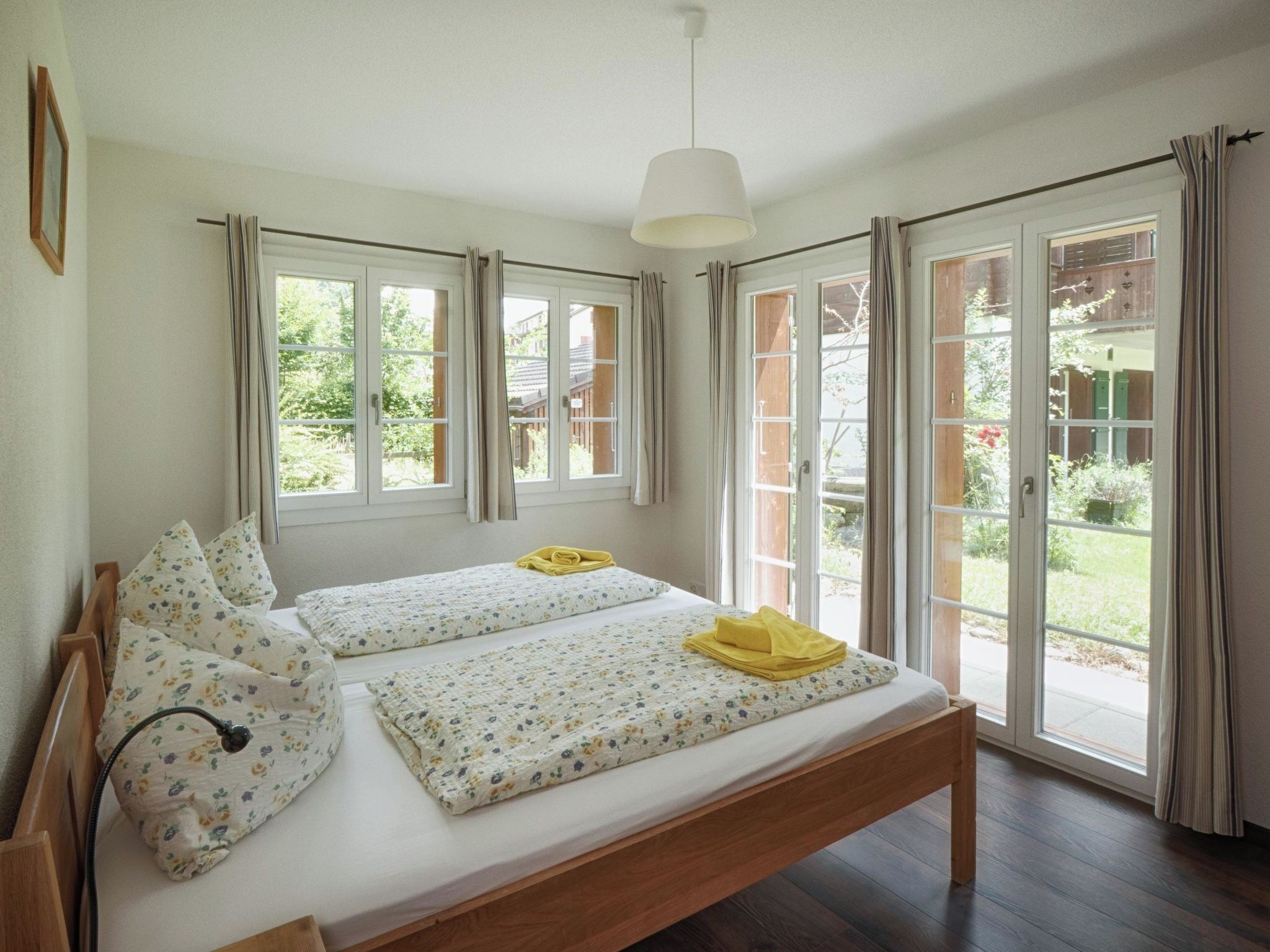 Un letto matrimoniale con lenzuola floreali e due cuscini, un comodino con una lampada, e vista su un giardino attraverso ampie finestre.