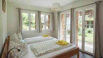 Un letto matrimoniale con lenzuola floreali e due cuscini, un comodino con una lampada, e vista su un giardino attraverso ampie finestre.