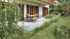En træbygning med en overdækket terrasse, et bord med stole, og en have med forskellige planter og blomster.
