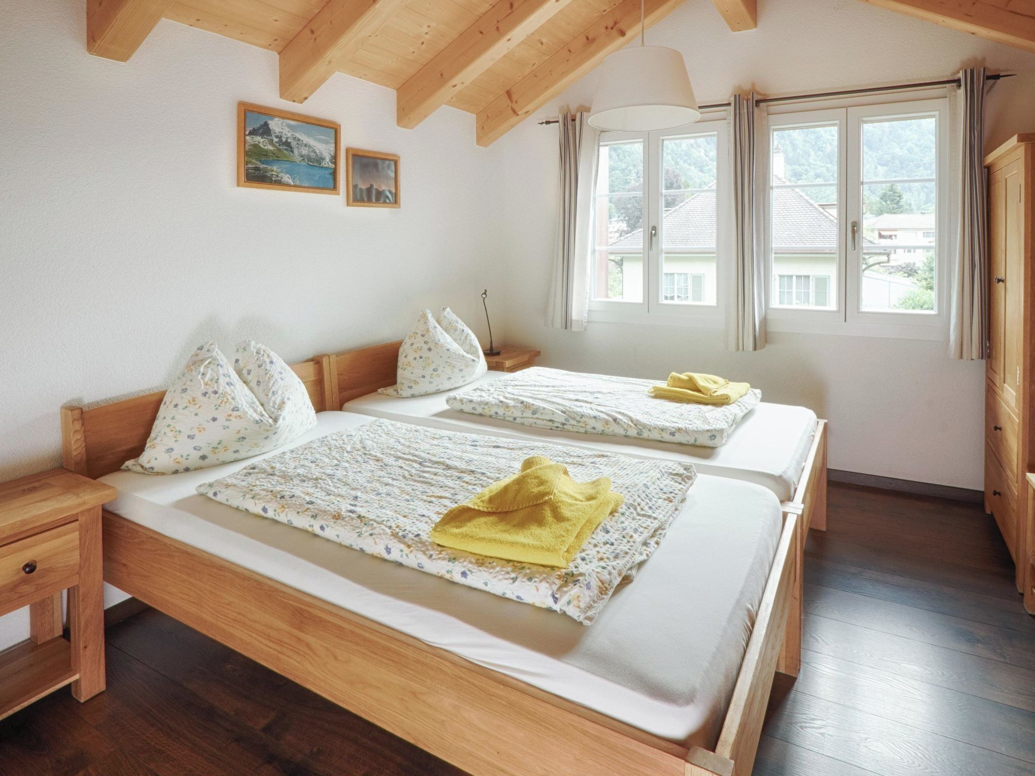Una stanza con due letti di legno, un comodino, una finestra con tende e un soffitto in legno.