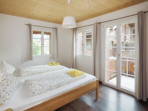 Une chambre avec un lit en bois, deux fenêtres et une vue sur une terrasse en bois à l’extérieur.