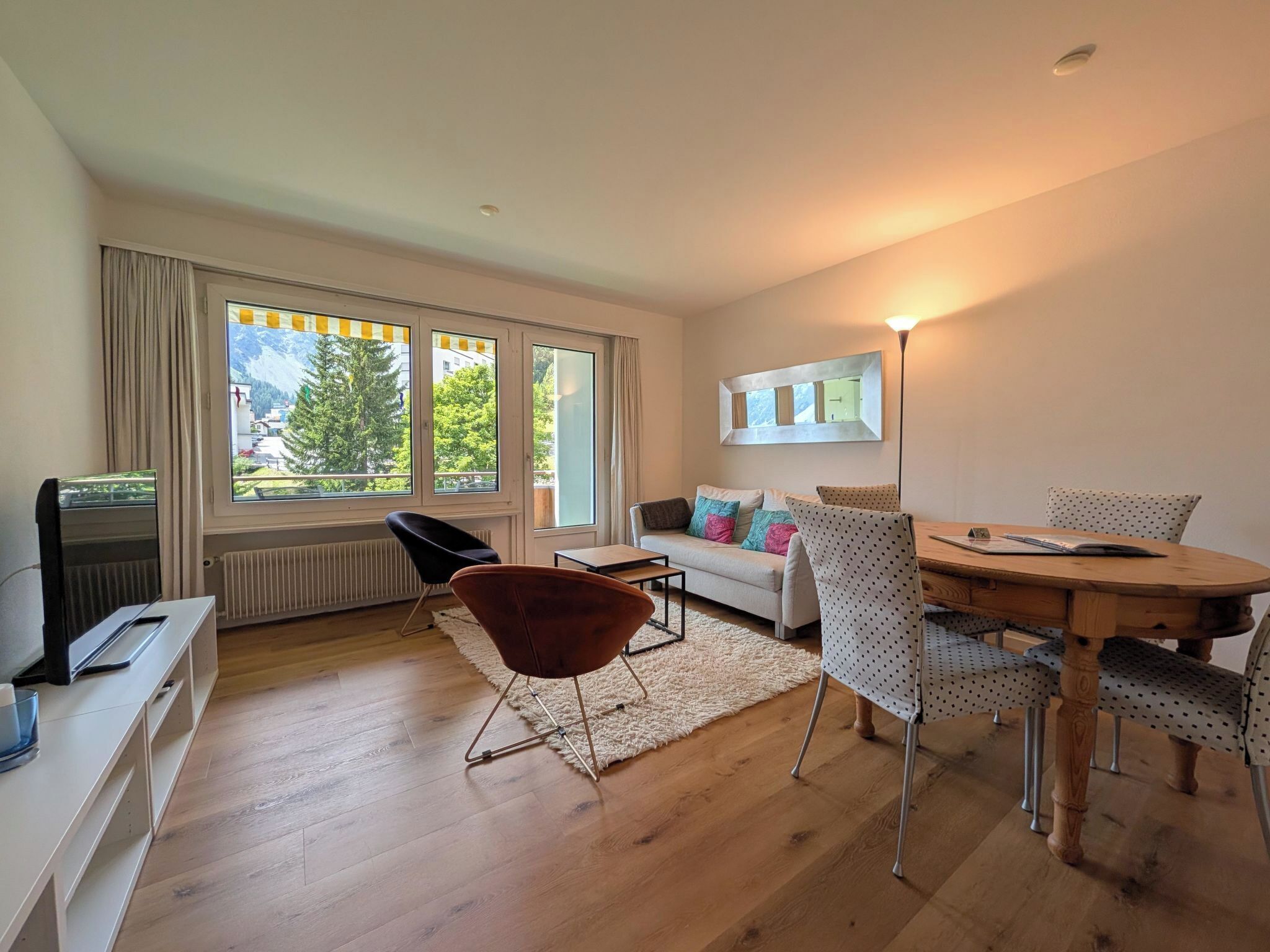 A modern living room with a sofa, dining table, chairs, a TV, and large windows with a view of greenery and mountains.
