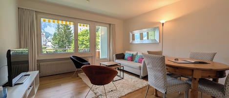 Ein modernes Wohnzimmer mit einer Couch, einem Esstisch, Stühlen, einem Fernseher und großen Fenstern mit Blick auf Grünflächen und Berge.