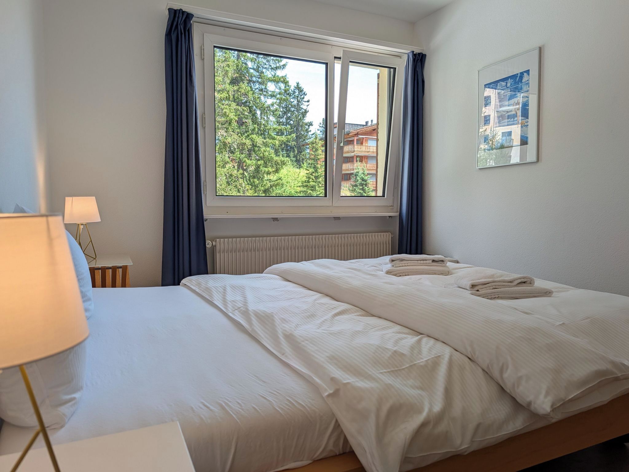 Habitación de hotel con una cama, ventana con cortinas, radiador, lámpara y un cuadro en la pared.