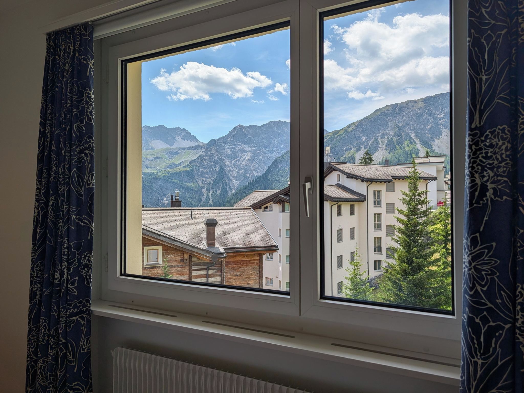 Una ventana con vistas a un paisaje montañoso, un edificio de techo de tejas y un árbol.