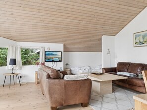 Ein modernes Wohnzimmer mit Holzboden, einem Flachbildfernseher, einem braunen Ledersofa, einem Holztisch und Blick ins Grüne.