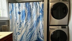 A laundry room with a double-door washer and dryer, a marble-patterned shower curtain, and a wooden floor. - Coastal Retreat Charming and Salty in St. Andrews/Panama City Florida (Panama City)