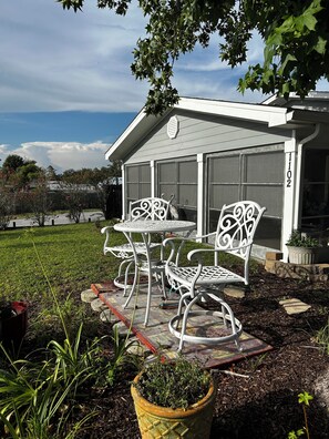 Eine Terrasse mit weißen Metallstühlen und einem Tisch, ein Blumentopf und im Hintergrund ein Haus mit einer Garagentür.