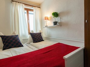 Une chambre à coucher avec un lit, des oreillers, une couverture rouge, une fenêtre avec des rideaux, une étagère fixée au mur avec une plante et une lampe.
