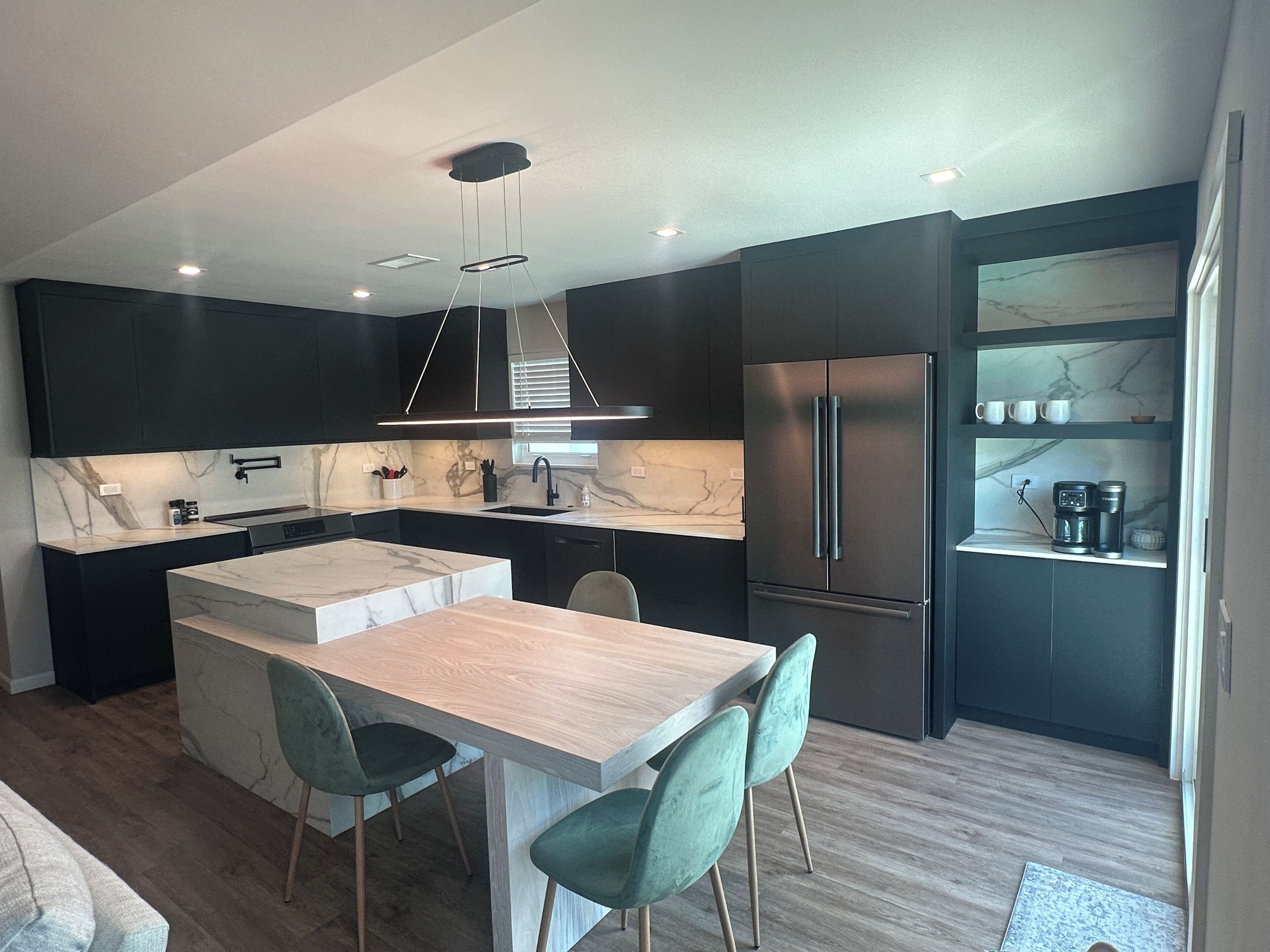Une cuisine moderne avec des meubles de couleur sombre, un pare-feu en marbre, un îlot central doté de chaises et un réfrigérateur encastré.