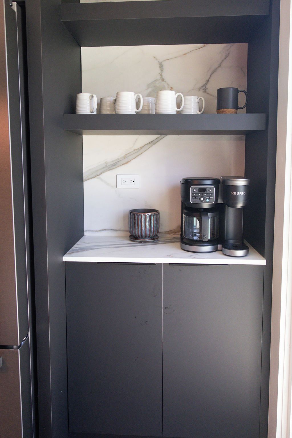A kitchen area with a marble countertop, a coffee maker, a kettle, and stacked white mugs on a shelf.