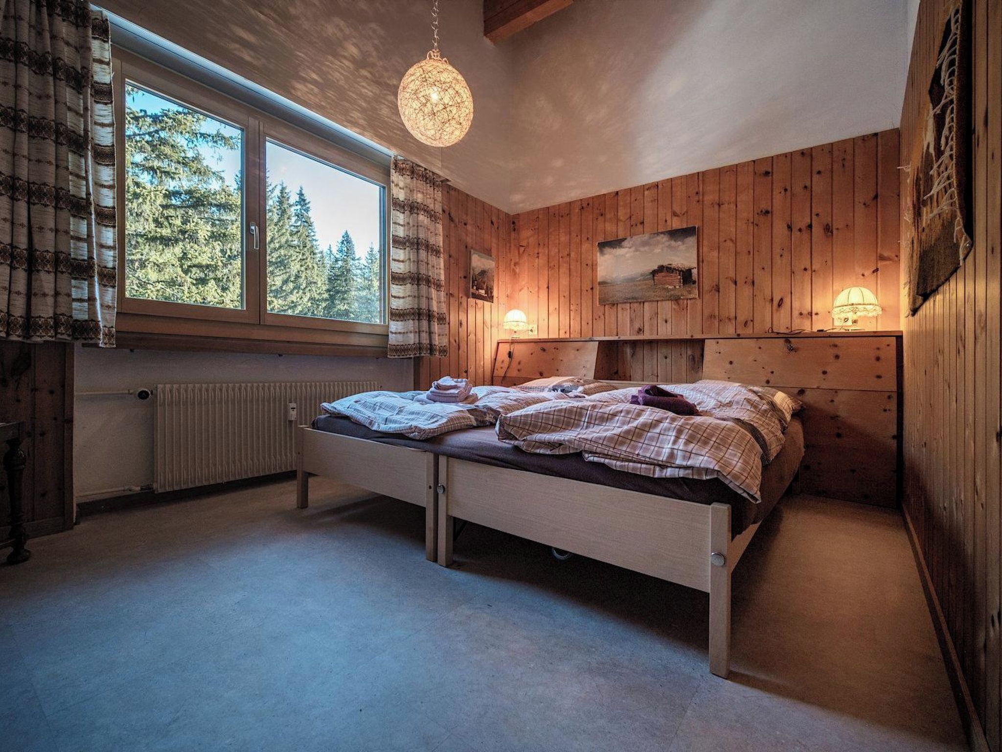 Een slaapkamer met houten vloer, een bed met wit frame, een raam met gordijnen, een hanglamp en een schilderij aan de muur.