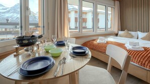 Ein Hotelzimmer mit Essbereich, einem Bett mit orangefarbener Decke und großen Fenstern mit Blick auf die Berge.