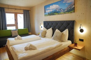 A double bed with white linens, a green sofa, wooden flooring, a painting of mountains, and a nightstand with a vase. - Benedikt – a cozy apartment steeped in history, perfect for unforgettable getaways (Gröbming)