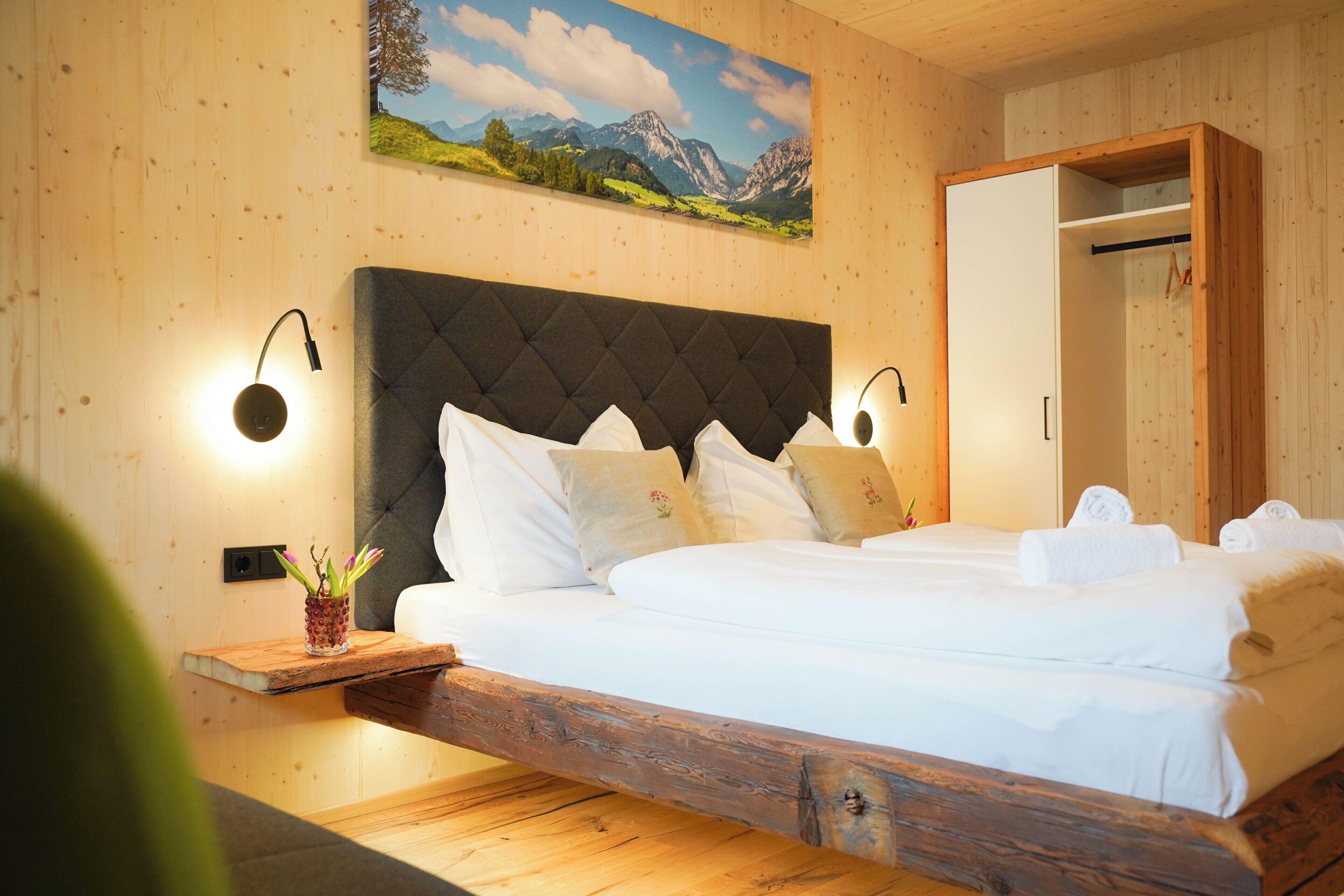 A bedroom with a bed, wooden headboard, bedside lamps, a painting, and a wooden nightstand with a plant.