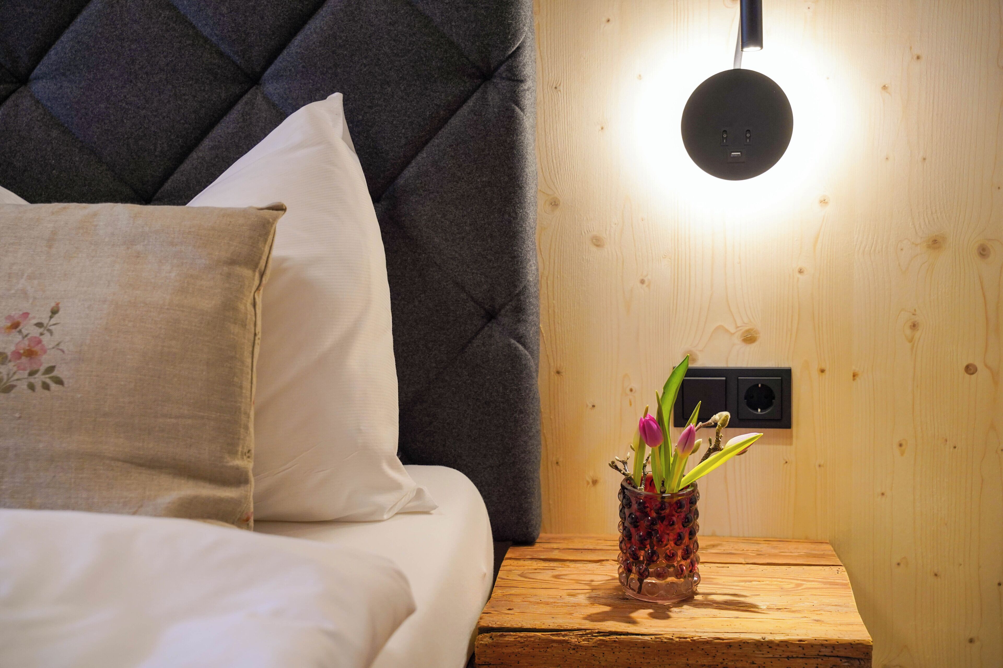 Un dormitorio con cama, almohadas, una mesita de noche con un florero y una lámpara de pared.