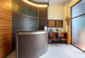 A reception desk with a logo, a wooden panel wall, a tiled floor, and a seating area with chairs and a vase of flowers. - Stylish Stay Near Malioboro (Yogyakarta)