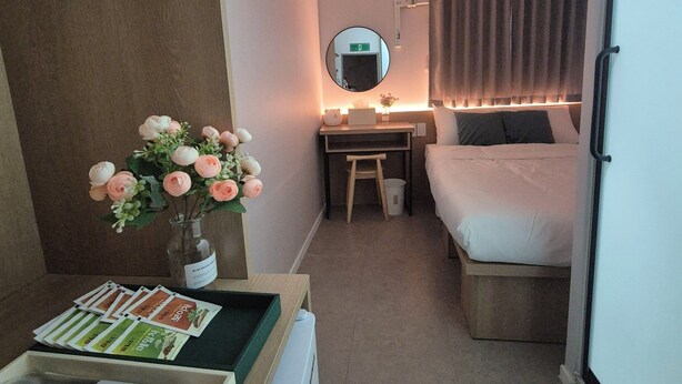 A hotel room with a bed, a desk with a chair, a mirror, a vase with flowers, and a tray with brochures. - Nana Hostel jonggak (Seoul)