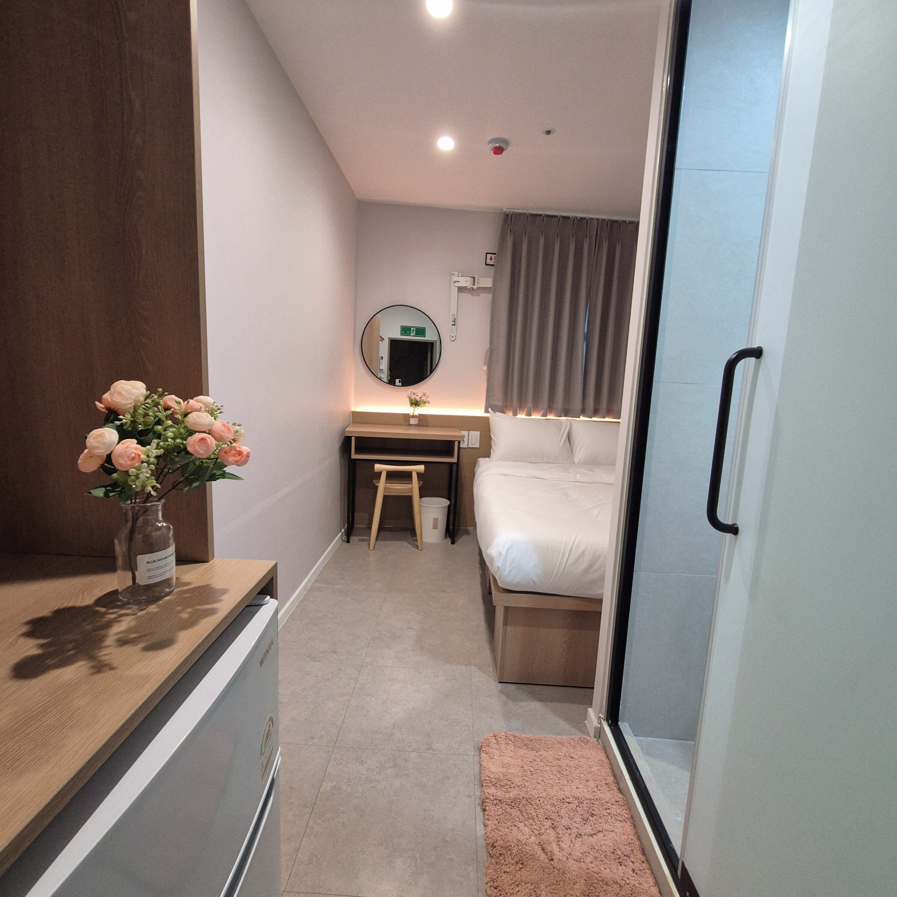 A compact hotel room with a bed, a desk, a chair, a small stool, a mirror, and a vase with flowers.