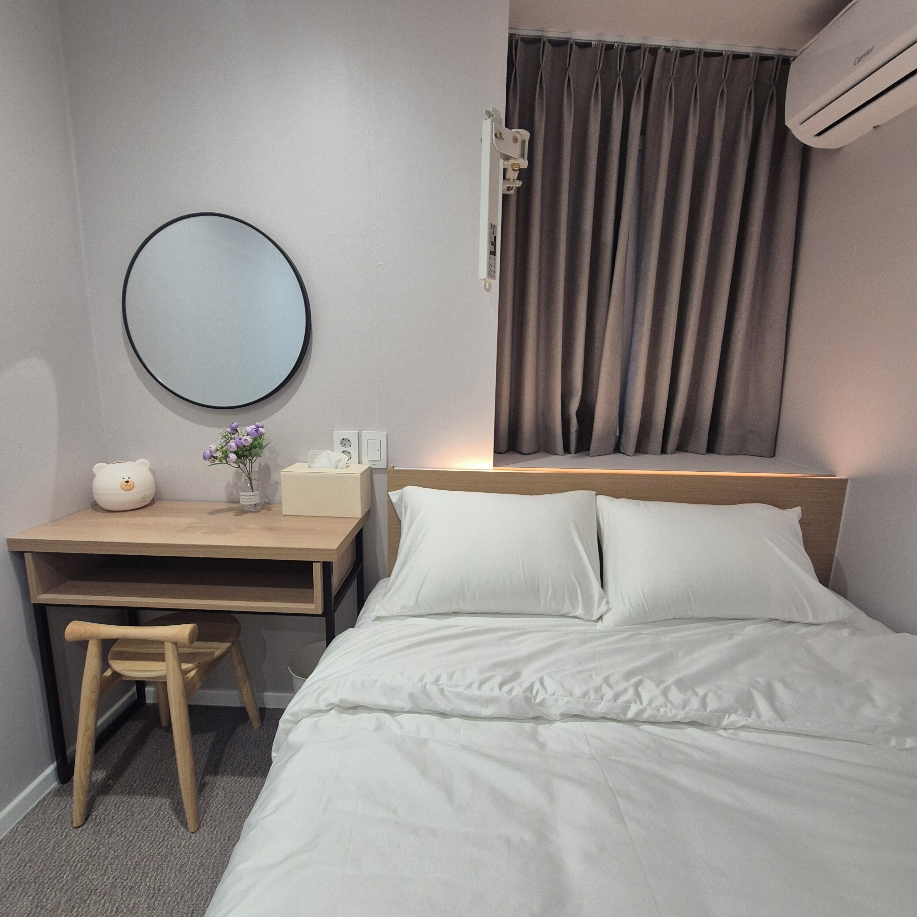 A hotel room with a bed, a desk with a chair, a round mirror, a bedside table with a lamp, and an air conditioning unit.