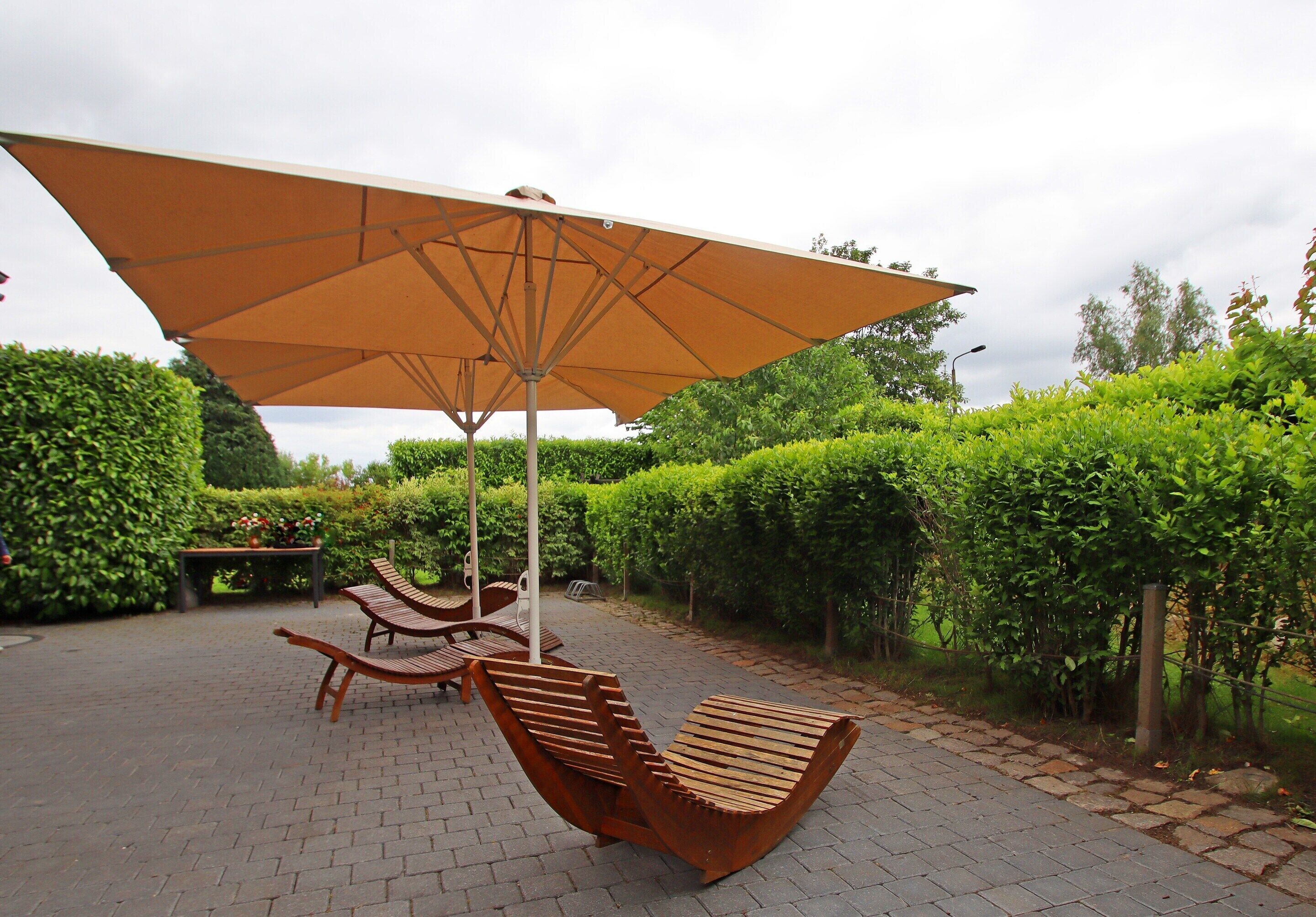 A patio area with two wooden lounge chairs and a large umbrella, surrounded by well-maintained hedges and a brick pathway.