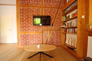 Ein Zimmer mit Backsteinwand, einem daran befestigten Flachbildfernseher, einem hölzernen Bücherregal und einem runden Holztisch.