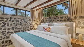 Un dormitorio con pared de piedra, una cama con ropa blanca, un plaid verde aguamarina y una mesita de noche con lámpara.