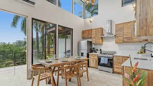 Una cocina moderna con armarios de madera, un comedor con mesa y sillas, y ventanas grandes que ofrecen vistas a palmeras.